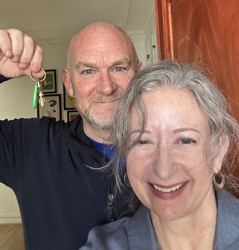 A smiling man holding a set of keys in his hand stands behind a smiling woman with gray hair. They are posing for a selfie in a well-lit indoor setting.